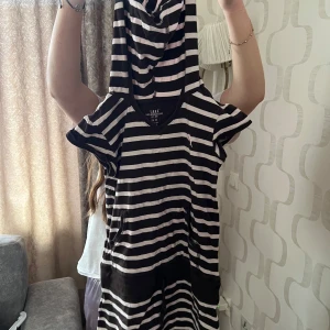 Svartvit randig klänning med huva - Säljer en svartvit randig klänning med korta ärmar och huva. Klänningen har v-ringning och är i t-shirtmaterial, perfekt för en avslappnad stil. Passar dig som gillar sportig och bekväm look.