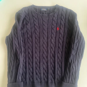 Mörkblå stickad tröja från Ralph Lauren - Säljer en klassisk mörkblå kabelstickad tröja från Ralph Lauren med rund halsringning och röd broderad logga på bröstet. Perfekt för dig som gillar tidlös stil och vill ha något både snyggt och bekvämt.