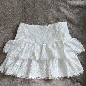 Vit volangkjol med broderade blommor - Superfin vit kjol med två volanger och broderade blommor. Kjolen har spetsdetaljer längs volangerna och är kort i modellen, storlek XL men sitter som en L, aldrig använd, prislapp kvar!