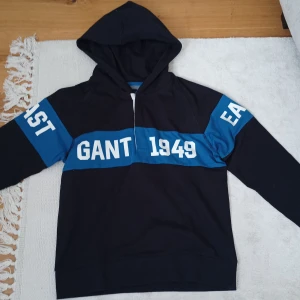 Svart och blå hoodie från GANT - Säljer en svart hoodie från GANT med blåa detaljer och vit text. På framsidan står det 'GANT 1949. Och på sidan av armarna står det EAST COAST' och på baksidan 'CONNECTICUT'. Tröjan har huva och knappar, perfekt för en sportig och avslappnad stil.