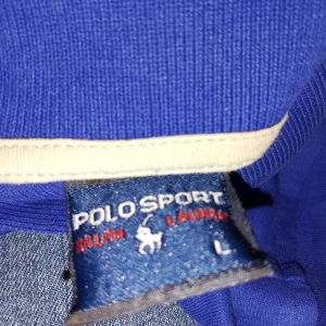 Blå pikétröja från Polo Sport Ralph Lauren - Säljer en klassisk blå pikétröja från Polo Sport Ralph Lauren med vit logga på bröstet och siffran 3 på ärmen. Tröjan har krage med vit kant och korta ärmar. Perfekt för en sportig och stilren look.