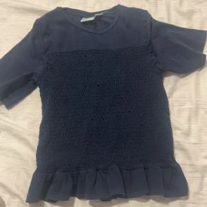 Mörkblå topp med volangkant. Använd ett få tal gånger - Säljer en mörkblå topp med korta ärmar och volangdetalj längst ner. Toppen har en smockad mittdel som ger en snygg struktur och en mjuk, rund halsringning. Perfekt för dig som gillar en söt och enkel stil.