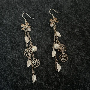 Långa silverfärgade örhängen med blommor och pärlor - Säljer ett par eleganta, långa örhängen i silverfärg med detaljer som blommor, löv och vita pärlor. Örhängena har flera kedjor som ger ett unikt och stilrent intryck. Perfekta för att lyfta din outfit!