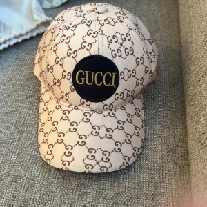 Beige keps med mönster - Säljer en beige keps med mönster i brunt och en svart patch med guldig text framtill. Kepsen har böjd skärm och justerbar passform. Bara att skriva vid frågor och priset kan såklart diskuteras!