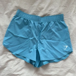 Blå shorts - Säljer dessa blåa träningsshorts från Gymshark i storlek S. Otroligt luftiga och väger ingenting! Perfekt till gymmet eller en varm sommardag! Har aldrig använt dessa då jag glömde skicka tillbaka! 