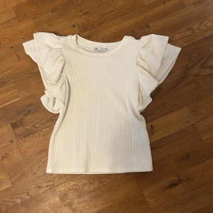 Vit ribbad topp med volangärmar från Zara - Supersöt vit topp från Zara med ribbat tyg och volangdetaljer på axlarna. Använd fåtal gånger!