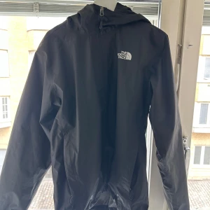 Svart vindjacka från The North Face - Säljer en svart vindjacka från The North Face med huva och dragkedja. Jackan har meshfoder på insidan och logga på bröstet. Perfekt för blåsiga dagar och enkel att packa ner i väskan.