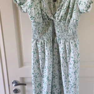 Vit och grön blommig klänning från SHEIN - Säljer en söt vit klänning från SHEIN med grönt blommönster. Klänningen har smock i midjan och omlott-ringning upptill. Perfekt för varma dagar med sina korta puffärmar och luftiga material. 