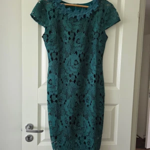 Spetsklänning med blommönster - Grön klänning med blommig spets och korta ärmar. Klänningen har rund halsringning dekorerad med spetsdetaljer och är figurnära i modellen
