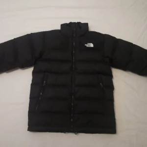 Svart dunjacka från The North Face - Säljer en svart dunjacka från The North Face med hög krage och dragkedja framtill. Jackan har två sidofickor med dragkedja och klassisk logga på bröstet. Perfekt för kalla dagar och enkel att matcha med olika outfits.fär man hänger upp jackan är sönder det går att fixa 