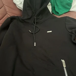 Svart hoodie från Diesel - Säljer en svart hoodie från Diesel med dragkedjedetalj vid fickan och justerbar huva med snören. Tröjan har en stilren look med en liten metallogo på bröstet. Perfekt för en avslappnad stil.