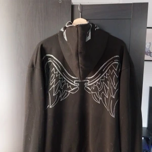 Svart rhinestone zip hoodie - Svart Rhinestone zip hoodie med dragkedja hela vägen upp. Rhinestone text på framsidan och wingar på baksidan även ansikte på luvan. Inga rhinestones saknas. Aldrig använd. (Kan finnas lite hund hår på tröjan)