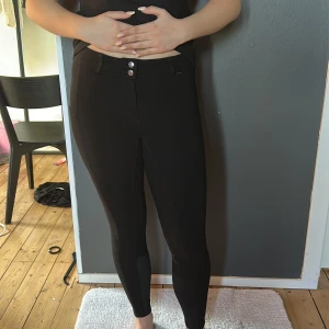 Svarta ridbyxor med hög midja - Säljer ett par svarta ridbyxor med hög midja, dubbla knappar framtill, snygga detaljer på insida lår samt fickor på utsida lår. Byxorna har tight passform och förstärkning på insidan av benen för extra komfort vid ridning. Perfekta för dig som vill ha stilrena och funktionella ridbyxor.