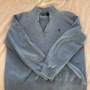 Ljusblå stickad tröja med dragkedja från Polo Ralph Lauren - Säljer en ljusblå stickad tröja från Polo Ralph Lauren med half zip. Tröjan har lång ärm, ribbade muddar och krage. Perfekt för sommaren eller vår