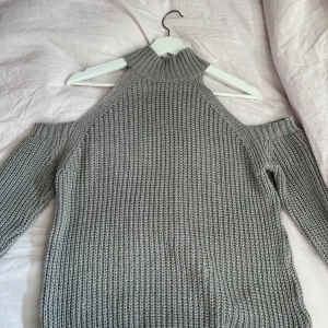 Grå stickad offshoulder tröja - Säljer en snygg grå stickad tröja med offshoulder-design och hög krage. Perfekt för kyliga dagar när du vill vara både varm och trendig. Tröjan har långa ärmar och en ribbad struktur som ger en skön passform. Det har en liten skada som jag fixat, men som inte syns!🩷