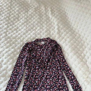 Blommig långärmad topp från Lindex - Säljer en söt långärmad topp från Lindex med ett blommigt mönster i rosa, lila och blå nyanser på en svart bakgrund. Perfekt för en färgglad look! 🌸säljer då den aldrig kommit till användning 