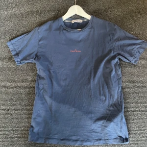 Blå t-shirt från Stone Island - Säljer en blå t-shirt från Stone Island med korta ärmar. Den har en subtil logga på framsidan och ett tryck på baksidan. Perfekt för en avslappnad stil. Passar bra till sommaren! Nypris 1900