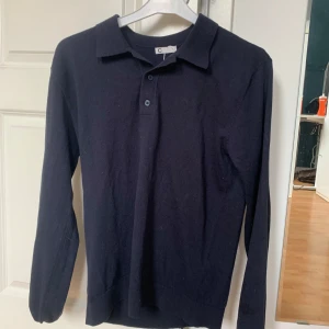 Svart långärmad pikétröja - Snygg svart långärmad pikétröja med knappar framtill. Perfekt för en stilren look. Tillverkad i ett mjukt material som ger en bekväm passform. Ett måste i garderoben för alla som gillar klassisk stil.