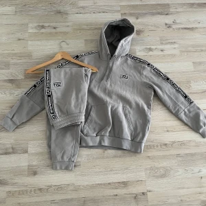 Grå hoodie och byxor från EA7 - Säljer en grå hoodie och matchande byxor från EA7. Setet har svarta detaljer med logotyp längs ärmar och ben. Hoodien har en klassisk känguruficka och byxorna har en bekväm resår i midjan. Perfekt för en avslappnad stil.