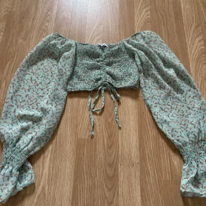 Blommig grön blus med puffärmar - Säljer en söt blommig blus i grönt med puffiga långa ärmar och knytning framtill. Blusen har ett elastiskt parti vid bysten och är perfekt för en somrig look.