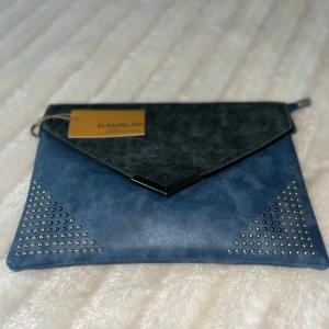 Blå och svart kuvertväska från Danblini - Snygg kuvertväska i blått och svart från Danblini. Väskan har en stilren design med nitar i hörnen och en praktisk dragkedja. Perfekt för att ge din outfit en extra touch.    ‼️Helt nya‼️