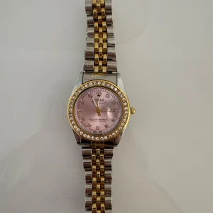  klocka med rosa urtavla - Säljer en elegant  klocka med en glittrig bezel och rosa urtavla. Klockan har en tvåfärgad länkarmband i guld och silver. Säljs då den inte kommit till användning.