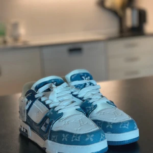 Blå och vita sneakers från Louis Vuitton - Snygga sneakers från Louis Vuitton i en fräsch kombination av blått och vitt. Skorna har ett distinkt mönster och tydlig logotyp på sulan. De är utrustade med snörning och har en rund tå. Perfekta för den som vill ha en stilren och lyxig look.