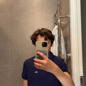 Hej jag seljer en mörkblå Lacoste t-shirt - Säljer en klassisk mörkblå pikétröja från Lacoste med den ikoniska krokodilloggan på bröstet. Tröjan har korta ärmar och en stilren design som passar perfekt till både vardag och mer uppklädda tillfällen.