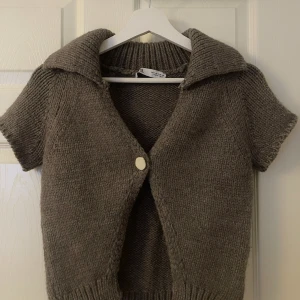 Beige stickad kofta från Zara - Säljer en mysig beige stickad kofta från Zara med korta ärmar och en stor krage. Koftan har en enkel knapp framtill och ribbade detaljer vid ärmslut och nederkant. Perfekt för lager-på-lager-stil!