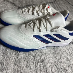 Copa Pure 2 - Snygga vita fotbollsskor från Adidas med blå och röda detaljer. Skorna har en blå sula med dobbar för konstgräs . Skorna är gjorda med läder vilket säkerställa mer skydd på planen. Ny pris 1600kr