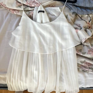 Vitt linne från Massimo dutti - Säljer ett elegant vitt linne med tunna axelband och volangdetaljer från Massimo dutti. Blusen har en luftig och lätt design med plisserad nederdel, perfekt för varma dagar. Passar både till jeans och kjol för en stilren look. 