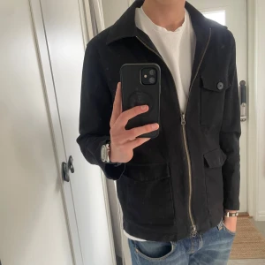 Svart jacka från John Henric - Snygg svart jacka från John Henric med dragkedja och knappar. Jackan har två bröstfickor och två fickor nedtill, alla med knappstängning. Perfekt för en stilren look.