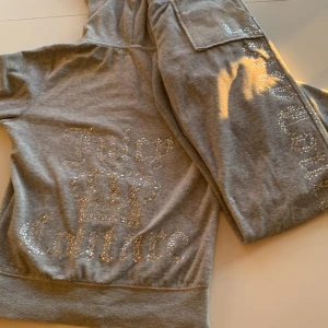 Gråt set från Juicy Couture - Säljer en snygg gråt set från Juicy Couture med glittrande stenar som bildar logotypen på ryggen och på byxorna. Hoodien har en dragkedja framtill och en bekväm passform. Perfekt för en avslappnad stil med en touch av glamour.En av knapparna saknas dock från ena bakfickan men inget som märks.🩶✨