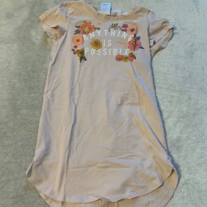 Beige t-shirtklänning med blommönster - Säljer en beige pjyamas klänning med texten 'Anything is Possible' och färgglada blommor på framsidan. Pjyamasen har korta ärmar och en rundad nederkant. Perfekt för en avslappnad stil.