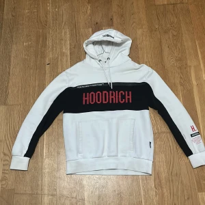 Hoodrich - Säljer en snygg vit och svart hoodie från Hoodrich med röd text på bröstet. Hoodien har en stor ficka framtill och justerbar huva med snören. Perfekt för en casual look.