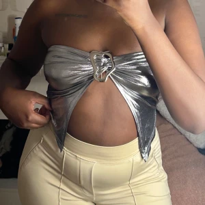 Silver axelbandslös tubtopp - Snygg och glittrig silverfärgad tubtopp med en unik metallringdetalj framtill. Perfekt för en utekväll eller fest. Toppen är axelbandslös och har en glansig finish som verkligen sticker ut 🩶