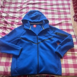 Blå hoodie från Nike - Säljer en snygg blå hoodie från Nike med dragkedja och huva. Jackan har en praktisk ficka med dragkedja på ärmen och svarta detaljer. Perfekt för en sportig look!