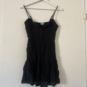 Svart axelbandslös klänning - Snygg svart klänning med smala axelband och knappar framtill. Perfekt för varma dagar med sin lätta och luftiga design. Den har en volangdetalj vid fållen som ger en extra touch av stil.