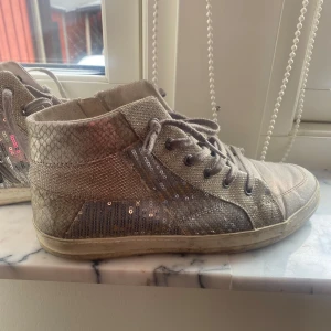 Glittriga sneakers med dragkedja - Snygga sneakers i beige med glittriga paljettdetaljer. Skorna har snörning och en dragkedja på sidan. 