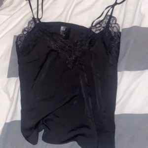 Svart linne med spetsdetaljer från H&M - Säljer ett elegant svart linne från H&M med tunna axelband och vacker spetsdetalj vid halsringningen. Perfekt för en stilren look. Passar bra till både jeans och kjol.