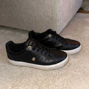 Tommy Hilfiger - Snygga svarta sneakers med gulddetaljer och snörning. Skorna har en vit sula och en diskret logga på sidan. Perfekta för en stilren look.