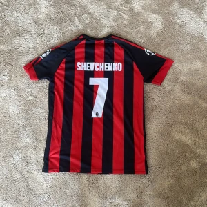 Shevchenko nummer 7, Ac Milan  - Säljer en klassisk röd och svart randig fotbollströja från Adidas med korta ärmar. Tröjan har tryck på både fram- och baksidan, inklusive ett stort nummer 7 och namn på baksidan. Perfekt för fotbollsfans! ⚽