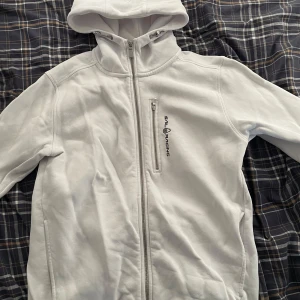 Vit zip från Sail Racing - Säljer en stilren vit zip hoodie från Sail Racing med dragkedja och huva. Hoodie’n har en broderad logotyp på bröstet och en praktisk bröstficka med dragkedja. Perfekt för en sportig look!