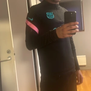 Barcelona träningströja - Säljer en svart långärmad träningströja från Nike med turkosa och rosa detaljer. Tröjan har en halv dragkedja framtill och klubbmärke på bröstet. Perfekt för fotbollsträning eller annan sportaktivitet.