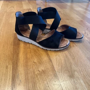 Svarta sandaler  - Snygga svarta sandaler med elastiska band och kilklack. De har en öppen tå och en bekväm sula. Perfekta för sommarens äventyr!