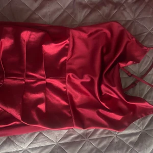 Röd axelbandslös klänning - En röd axelbandslös klänning i satin med en elegant passform. Perfekt för speciella tillfällen. Klänningen har en smickrande design med veckade detaljer framtill.Passar även 38