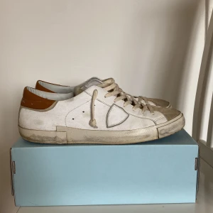 Vita sneakers från Philippe Model  - Vita  sneakers från Philippe Model med bruna detaljer och snörning. Skorna har en rund tå och är tillverkade i en kombination av skinn och mocka. Perfekta för en stilren look. Skorna är i storlek 47 men passar 46. Finns små defekter som lätt går att få bort med rätt tillbehör. Priset är inte skrivet i sten.