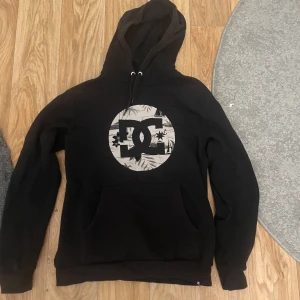 Svart hoodie från DC - Säljer en svart hoodie från DC med en stor logga på bröstet. Hoodien har en klassisk design med känguruficka och justerbar huva. Perfekt för en avslappnad stil.