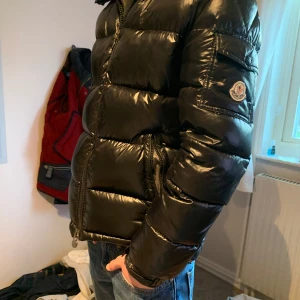 Svart pufferjacka från Moncler - Säljer en snygg svart pufferjacka från Moncler. Jackan har en glansig finish och är utrustad med dragkedja framtill. Den har långa ärmar och en praktisk huva för extra skydd mot väder och vind. Perfekt för kyligare dagar!