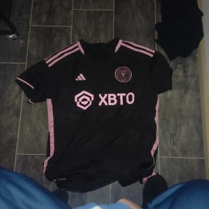 Svart fotbollströja messi miami  - Säljer en svart fotbollströja från Adidas med rosa detaljer och korta ärmar. Tröjan har ett tryck med nummer 10 och text på baksidan. Perfekt för fotbollsfans som vill ha en stilren look. De är messi och laget miami de är storlek L